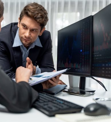 Two business traders discussing value business statistic graph company on paper report analyzing with dynamic stock exchange market currency real time website surround pc screens at office. Postulate.